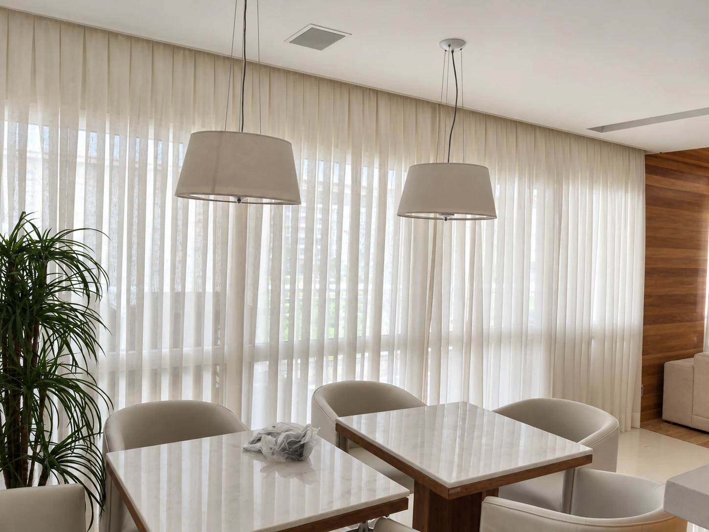 Cortinas claras de tecido em uma sala de jantar com duas mesas, cadeiras e luminárias suspensas.